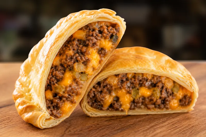 Empanada Cheeseburger de Las Tapas abierta al medio sobre una tabla de madera, bien rellena de carne picada con cheddar fundido, cebolla y pepinillo, con una gota de cheddar caliente cayendo del corte; detrás, un local de noche desenfocado